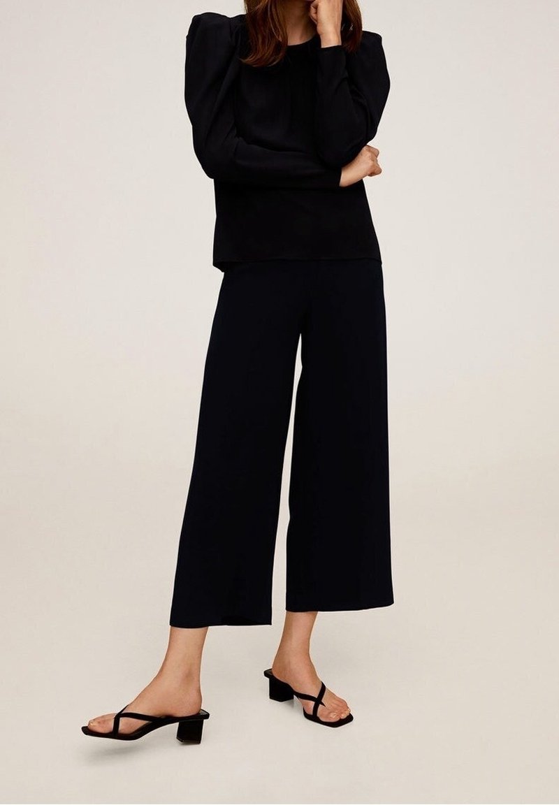 Černý dlouhý rukáv top s vycpanými rameny, spárovaný s černými širokými kalhotami a černými nazouvacími sandály na podpatku. Hladká látka, minimalistický design.