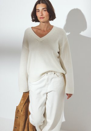 Cream v-neck sweater with long sleeves, paired with white high-waisted trousers. A tan suede jacket is held in the left hand.