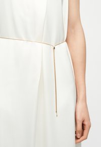 Ceinture fine en chaîne dorée attachée autour de la taille d'une robe blanche, avec la chaîne pendante d'un côté et une main reposant sur le côté.
