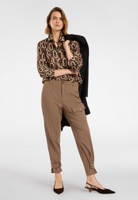 Patterned brown blouse with geometric design, beige tapered pants with side pocket and cuffs, black blazer draped over arm, brown heels.