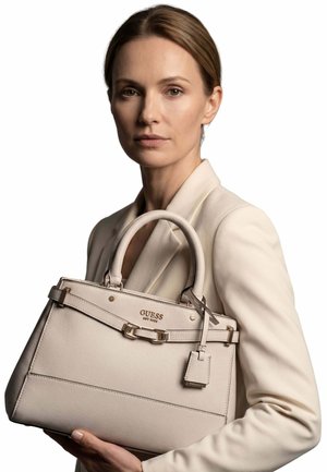 Femme en blazer beige tenant un sac à main Guess beige avec poignées supérieures et dorures sur fond blanc.