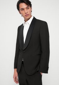 Homme portant une veste de smoking noire avec des revers en satin et une chemise blanche, regardant légèrement sur le côté contre un fond uni.