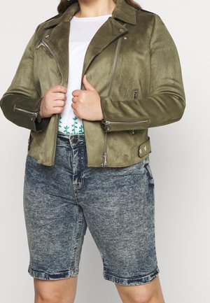 Person trägt eine olivgrüne Wildlederjacke mit silbernen Reißverschlüssen und Acid-Wash-Jeansshorts und hält den Jackenkragen mit beiden Händen.