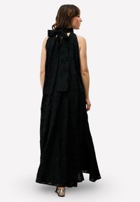 Zwarte halter maxi jurk met een textuurachtig bloemmotief, voorzien van een grote strik bij de hals en een gespleten rok; gedragen met zilveren schoenen.