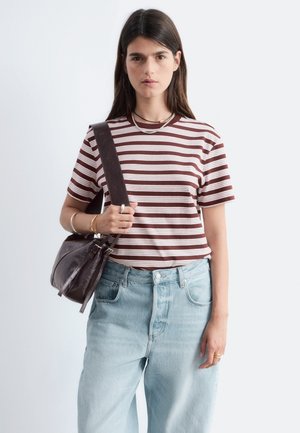 Mujer con cabello largo y oscuro, vestida con una camisa de rayas marrón y blanca, vaqueros de tiro alto azul claro, sosteniendo un bolso de hombro de cuero marrón oscuro.