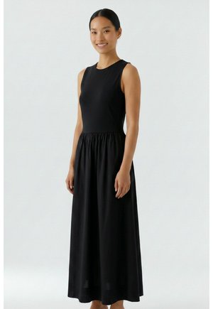 Femme debout et souriante, portant une robe noire sans manches, longue jusqu'aux chevilles, à corsage ajusté et jupe froncée, sur fond uni.