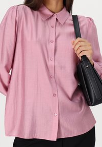 Blouse rose, en soie, avec un col, des manches longues et de légères pinces aux épaules. Elle présente des boutons assortis et une coupe ample.