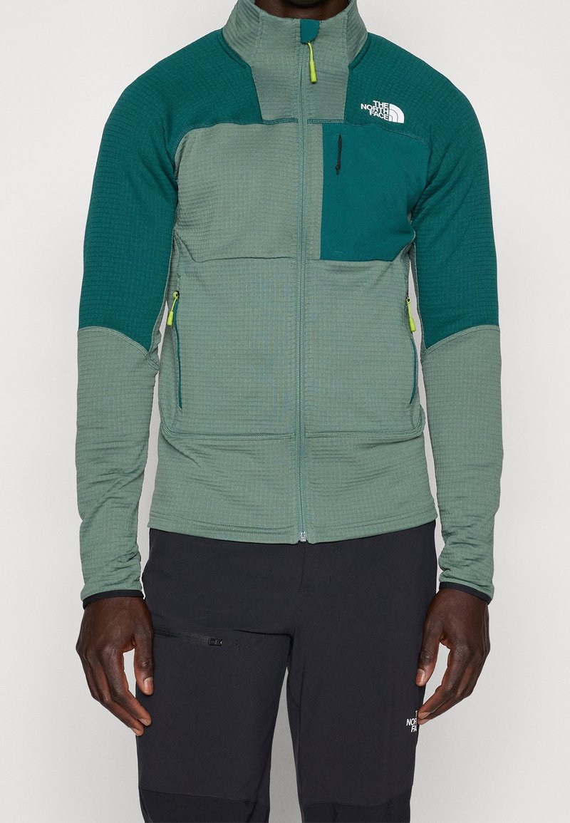 Homme portant une veste polaire verte zippée avec des manches et une poche poitrine vert foncé, associée à un pantalon noir.