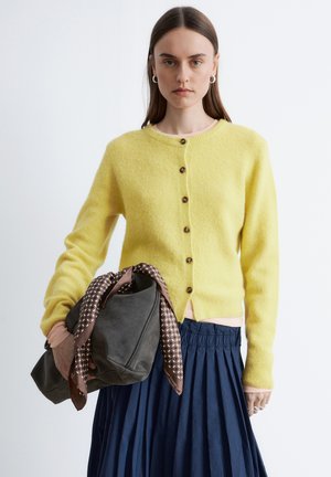 Kobieta o prostych brązowych włosach nosi żółty kardigan na guziki, granatową plisowaną spódnicę, trzyma szarą zamszową torbę z wzorzystą brązową chustą.