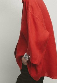 Chemise ample en lin rouge à manches courtes avec une poche, associée à un pantalon noir et des bracelets argentés au poignet.