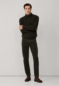 Pull à col roulé en maille torsadée vert foncé, associé à un pantalon ajusté vert foncé et des bottines en daim marron, sur un fond neutre.