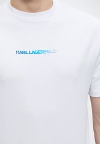 Άσπρο μπλουζάκι με το κείμενο «KARL LAGERFELD» σε διαβάθμιση αποχρώσεων του μπλε στο ύψος του στήθους, που φοριέται από άτομο κομμένο στο λαιμό και το χέρι.