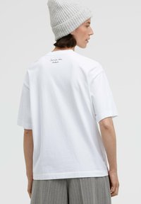 White cotton t-shirt with short sleeves, relaxed fit, and a small print on the back that reads "Summer the label." Textured gray knit beanie worn.