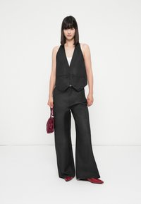 Mulher com cabelo liso e escuro, a usar um colete preto sem mangas, calças largas pretas, sapatos vermelhos e segurando uma pequena bolsa vermelha, contra um fundo branco.