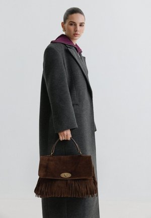 Femme portant un manteau gris foncé et des boucles d'oreilles dorées, tenant un sac à main en daim marron avec des franges et un fermoir, sur un fond clair uni.