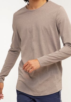 Homme portant une chemise à manches longues brun clair et un pantalon bleu marine, positionné avec une main près de la taille et l'autre légèrement étendue.