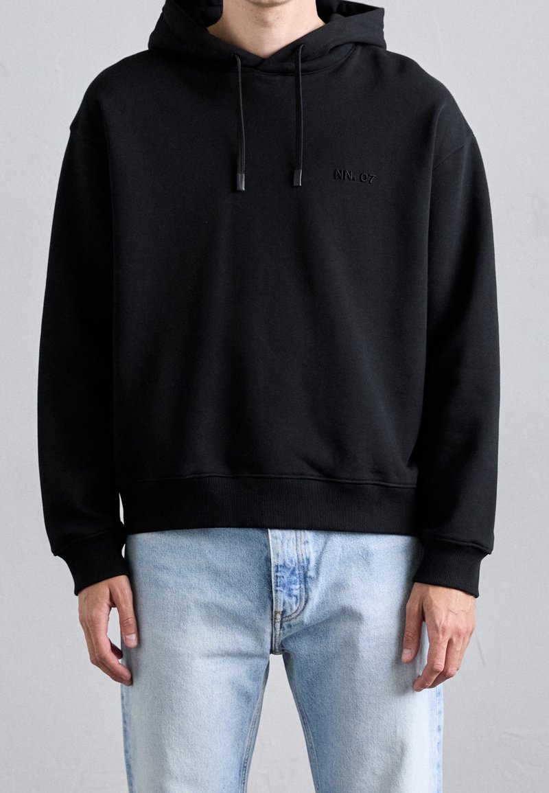 Homme portant un sweat à capuche noir avec un petit logo brodé "NN.07" sur la poitrine et un jean bleu clair, debout devant un fond gris.