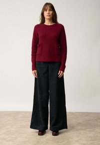 Bordeaux geribbelde trui met twee voorzakken, gecombineerd met brede zwarte corduroypantalons. De trui heeft lange mouwen en een ronde halslijn.