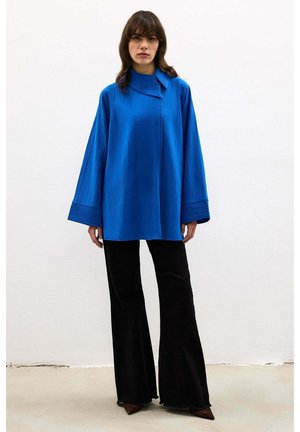 Vrouw staande, gekleed in een loszittende, langarmige blauwe top met brede manchetten en zwarte wijde broekspijpen, tegen een effen witte muur.