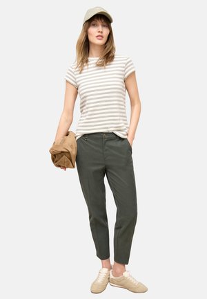 Femme portant un t-shirt beige à rayures, un pantalon vert olive, des baskets beiges et une casquette beige, tenant une veste marron pliée dans une main.