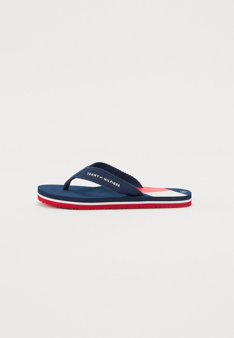 Navy flip-flop sandal with red and white striped sole. Textured fabric strap features "TOMMY HILFIGER" logo. Lightweight and flexible design.