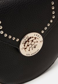 Schwarze Lederhandtasche mit rundem goldfarbenem Logoverschluss, strukturierter Oberfläche und dekorativen silbernen Nieten entlang des Randes.