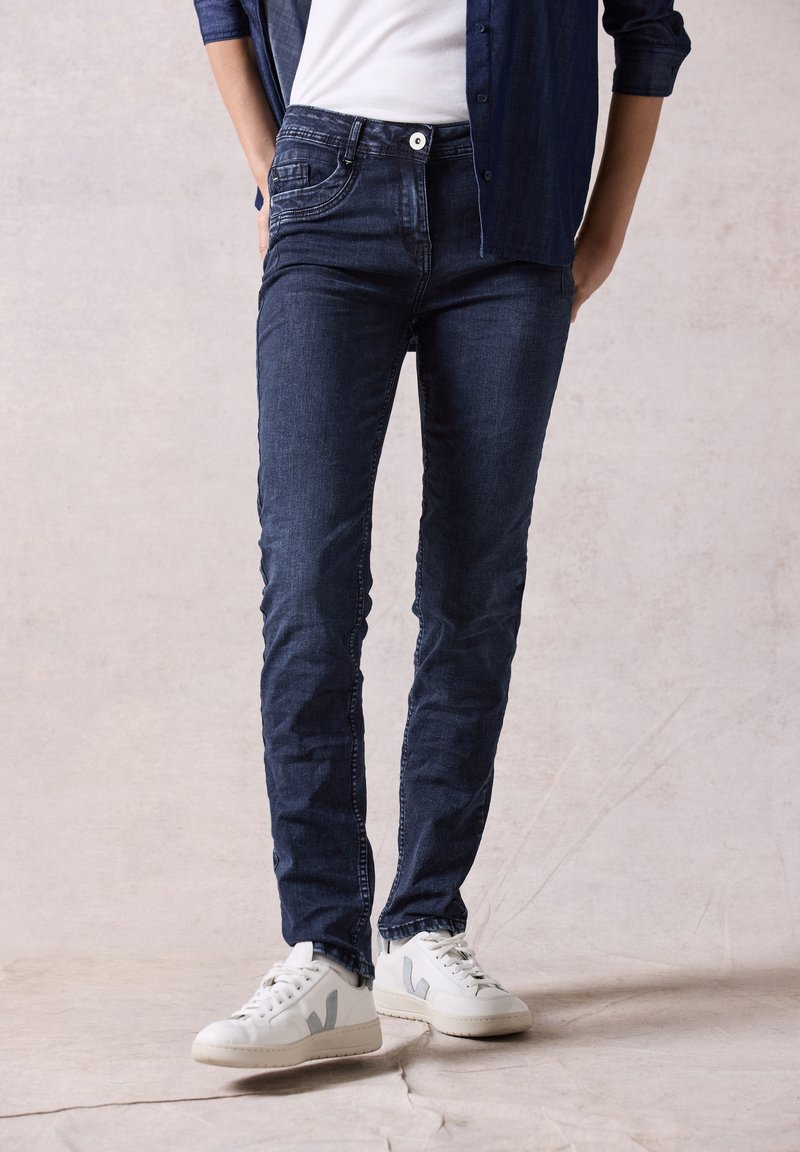 Jean slim fit bleu foncé avec fermeture à bouton et cinq poches, assorti à des baskets blanches avec des accents gris.