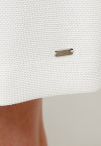 Gros plan sur un tissu blanc texturé avec une petite étiquette rectangulaire argentée marquée "Hilfiger" près du bord sur un fond de peau bronzée.