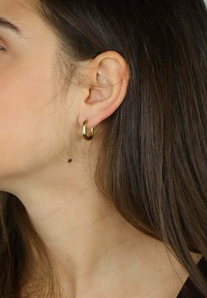 Złota kolczyk koło o gładkim wykończeniu, małym rozmiarze i okrągłym kształcie, noszony na płatku ucha na tle średnio brązowych włosów.