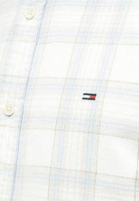 Cămașă albă în carouri cu linii bleu deschis și gri; are închidere cu nasturi și un mic logo brodat în roșu, alb și albastru pe piept.