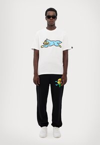 Hvid t-shirt med blåt tegneseriekat-design, parret med sorte bukser med et farverigt is-grafik. Båret med solbriller og hvide sneakers.