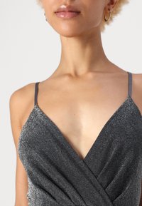 Λαμπερό μαύρο φόρεμα με λεπτές τιράντες, που διαθέτει βαθύ V-ντεκολτέ και πτυχώσεις. Το ύφασμα έχει υφή με λαμπερή φινέτσα.