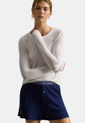Ung kvinde iført en langærmet hvid skjorte og mørkeblå Ralph Lauren-shorts med knappelukning står med armene over kors.