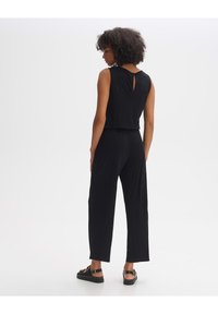 Schwarzer ärmelloser Jumpsuit mit einem Schlüssellochausschnitt am Rücken, lockerer Passform, geraden Beinen und weicher Textur, kombiniert mit schwarzen Sandalen.