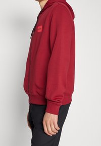 Červená mikina na zip s kapucí, vyrobená z měkkého materiálu, s logem na přední straně, žebrovanými manžetami a lemem. Nositá přes černé kalhoty.