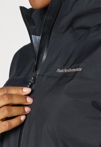 Black windbreaker jacket with zip closure; features PeakPerformance logo embroidery; smooth texture, high collar, and adjustable cuffs.