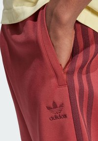 Czerwone/spodenki sportowe Adidas o gładkiej fakturze. Posiada boczne kieszenie, trzy ciemniejsze pionowe paski oraz haftowane logo na nodze.