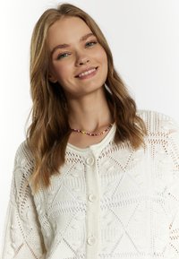 Vit stickad cardigan med ett geometriskt mönster, med stora knappar och en öppen väv. Färgglad pärloker gör detaljen runt halsen.