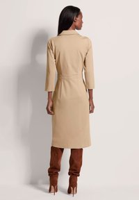 Beige Trenchcoat-Kleid mit Kragen, langen Ärmeln und taillierter Passform. Das Modell trägt braune Wildleder-Stiefel bis zum Oberschenkel mit einem Stiletto-Absatz.