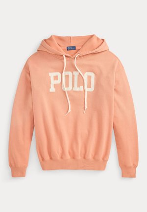Kobieta stoi, nosi morelową bluzę z kapturem "Polo", oliwkowe spodnie, brązowe sandały, trzyma dużą beżową torbę na ramię, jedną ręką w kieszeni.