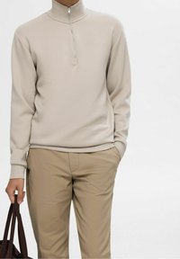 Sweatshirt beige zippé avec col haut, manches longues et texture lisse, associé à un pantalon kaki. Main tenant un sac fourre-tout marron.