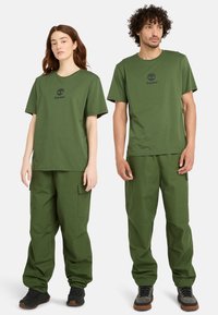 Gröna bomullströjor och cargo shorts, med en logotyp på bröstet. Löst snitt med sidofickor i matchande tyg. Svart fotbeklädnad.