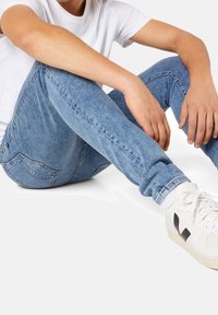 Jean en denim bleu clair à coupe ajustée, avec un léger effet délavé et deux poches arrière, associé à des baskets blanches et un t-shirt blanc uni.