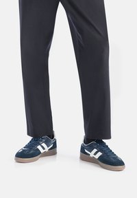 Granatowe sneakersy wykonane z zamszu i siateczki, z białymi akcentami, teksturowaną gumową podeszwą oraz niskoprofilowym designem, w zestawieniu z ciemnymi spodniami.