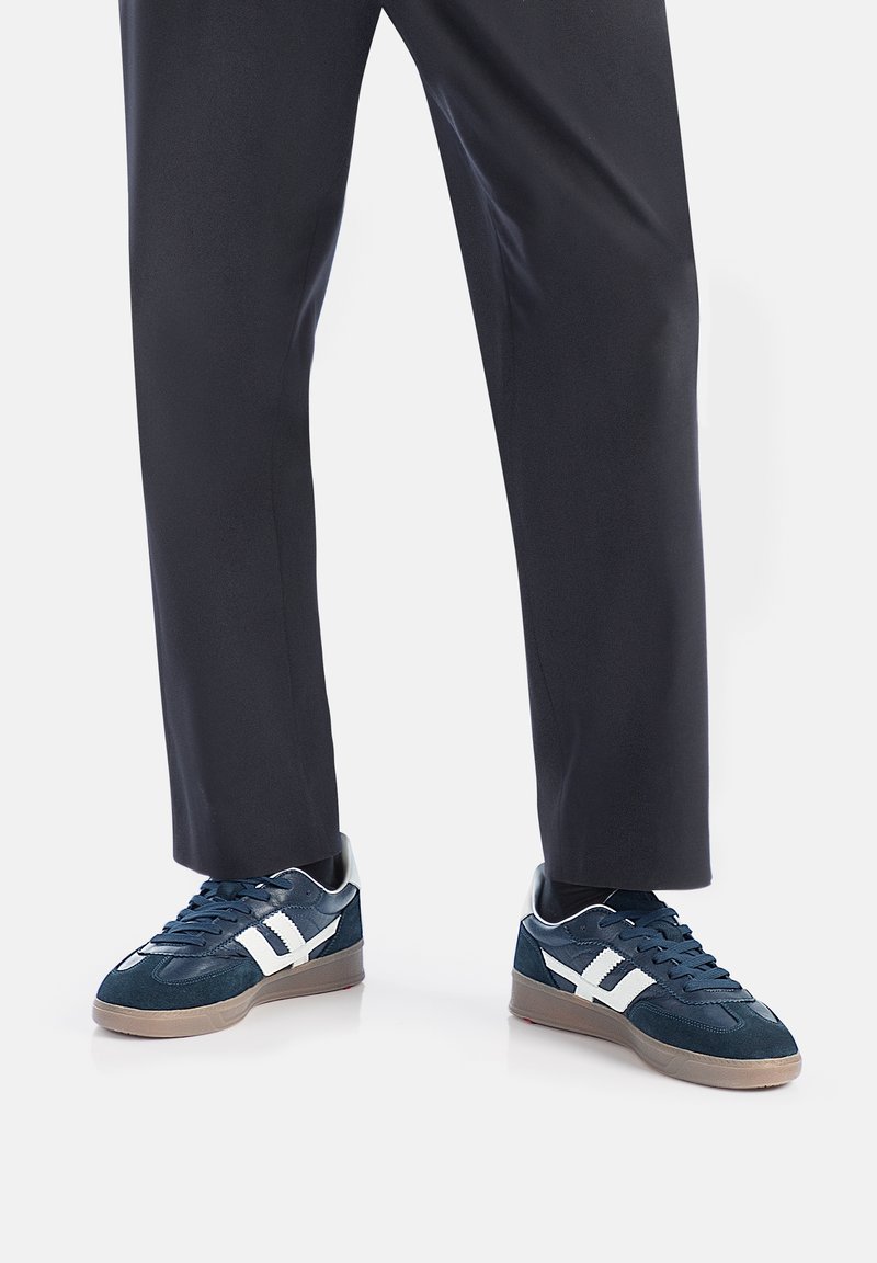 Granatowe sneakersy wykonane z zamszu i siateczki, z białymi akcentami, teksturowaną gumową podeszwą oraz niskoprofilowym designem, w zestawieniu z ciemnymi spodniami.