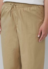Beige, leichte Hose mit elastischem Bund und Kordelzug, die seitliche Taschen und eine glatte Textur aufweist. Nahaufnahme von Taille und Hand.