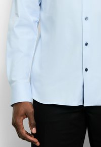 Hellblaues Langarmhemd mit strukturiertem Muster, ausgestattet mit dunklen Knöpfen, einem Kragen und einem geraden Saum. Getragen mit schwarzen Hosen.