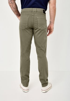 Mann in olivgrüner Hose, marineblauem Hemd und weißen Turnschuhen, der mit sichtbaren tätowierten Armen von hinten vor einem schlichten Hintergrund steht.