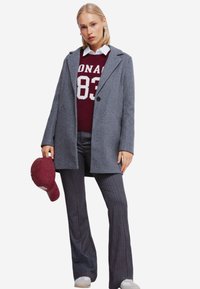 Grå uldfrakke med slagkrave, båret over en burgundy sweater med hvid tekst. Stratet grå bukser og en burgundy kasket fuldender outfittet.