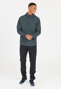 Leichte, dunkelgrüne Jacke mit durchgehendem Reißverschluss, elastischen Bündchen und zwei frontalen Taschen. Schwarze Hosen und blaue Sportschuhe runden das Outfit ab.
